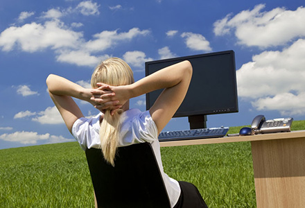 Woman sitting at an office desk with a computer out in a field with a blue sky