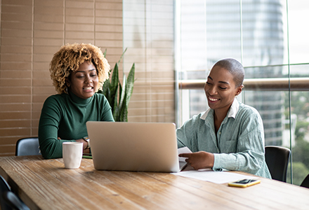 two women sitting at a large table looking at a laptop and smiling
